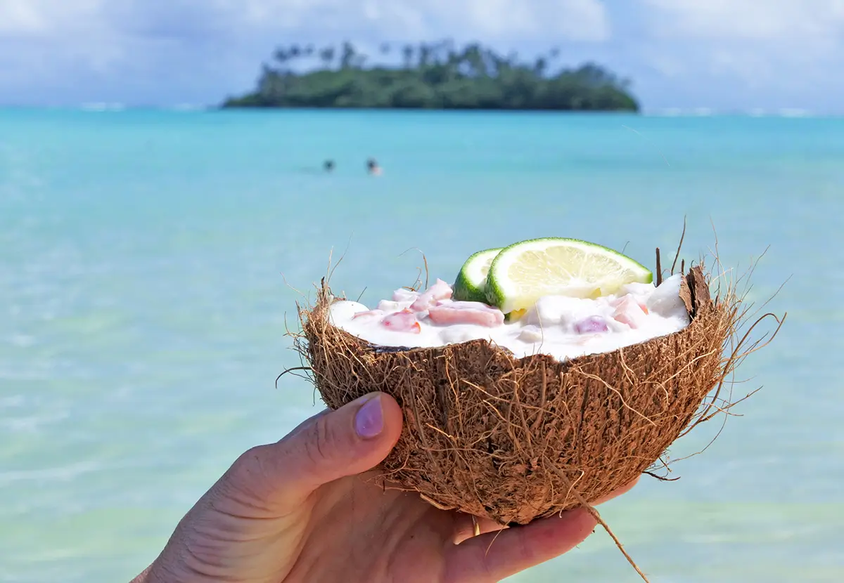 Woman holds Ceviche Dish served in a coconut shell against a islet in Muri lagoon Rarotonga